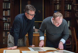 Two men looking at texts laid out on a wooden table in a library reading room