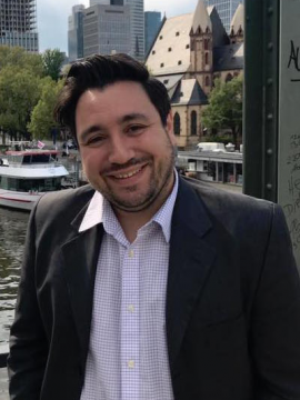 Chris Mustazza in a white shirt and black coat standing in front of a city scape.