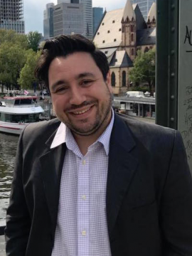 Chris Mustazza in a white shirt and black coat standing in front of a city scape. 