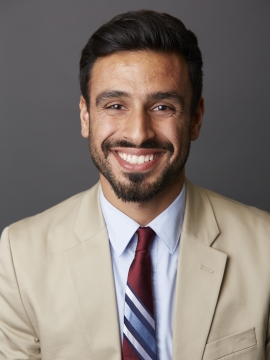 Dr. Ali M. Rahman in a beige blazer and a striped tie, seated in front of a gray background. 